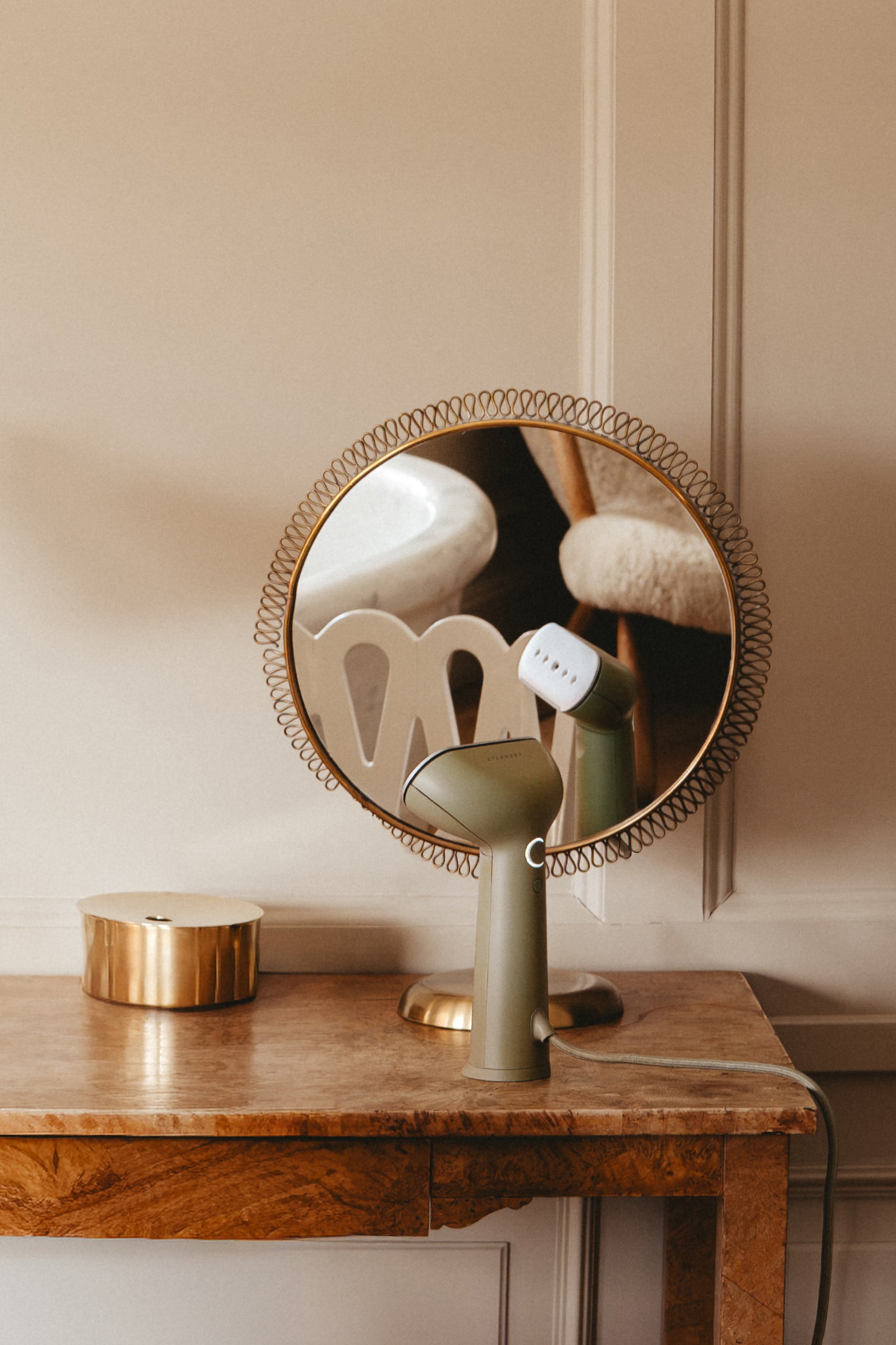 Green steamer in front of a round mirror with decorative frame on a wooden surface against a beige wall.