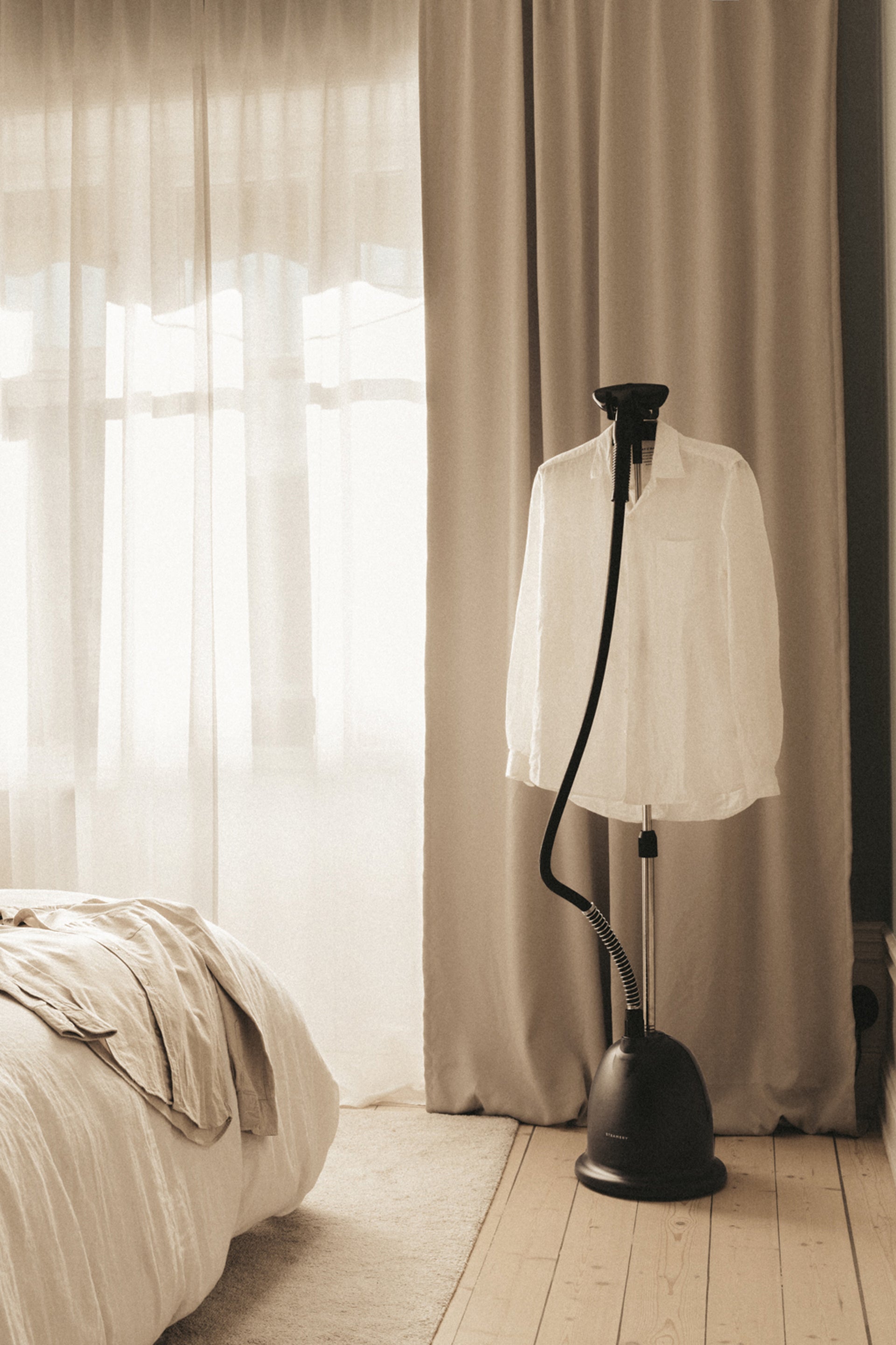 White shirt on a black floor steamer in a bedroom with curtains and bed in the background