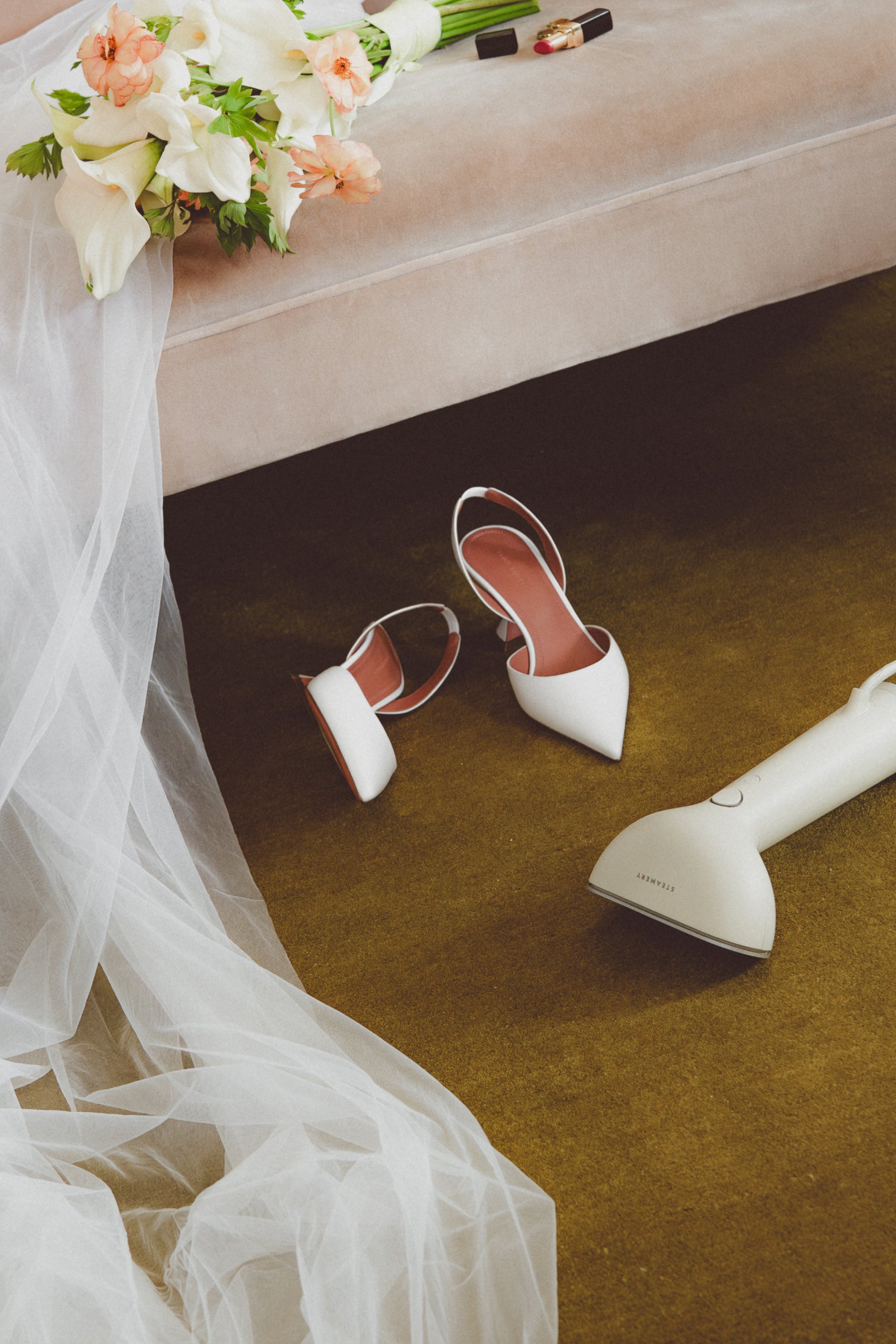 White high-heeled shoes and a white garment steamer on a carpeted floor with a wedding dress and bouquet in the background.