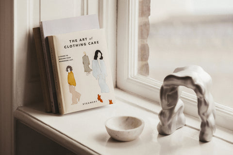 Book titled 'The Art of Clothing Care' on a windowsill with a small sculpture and bowl.