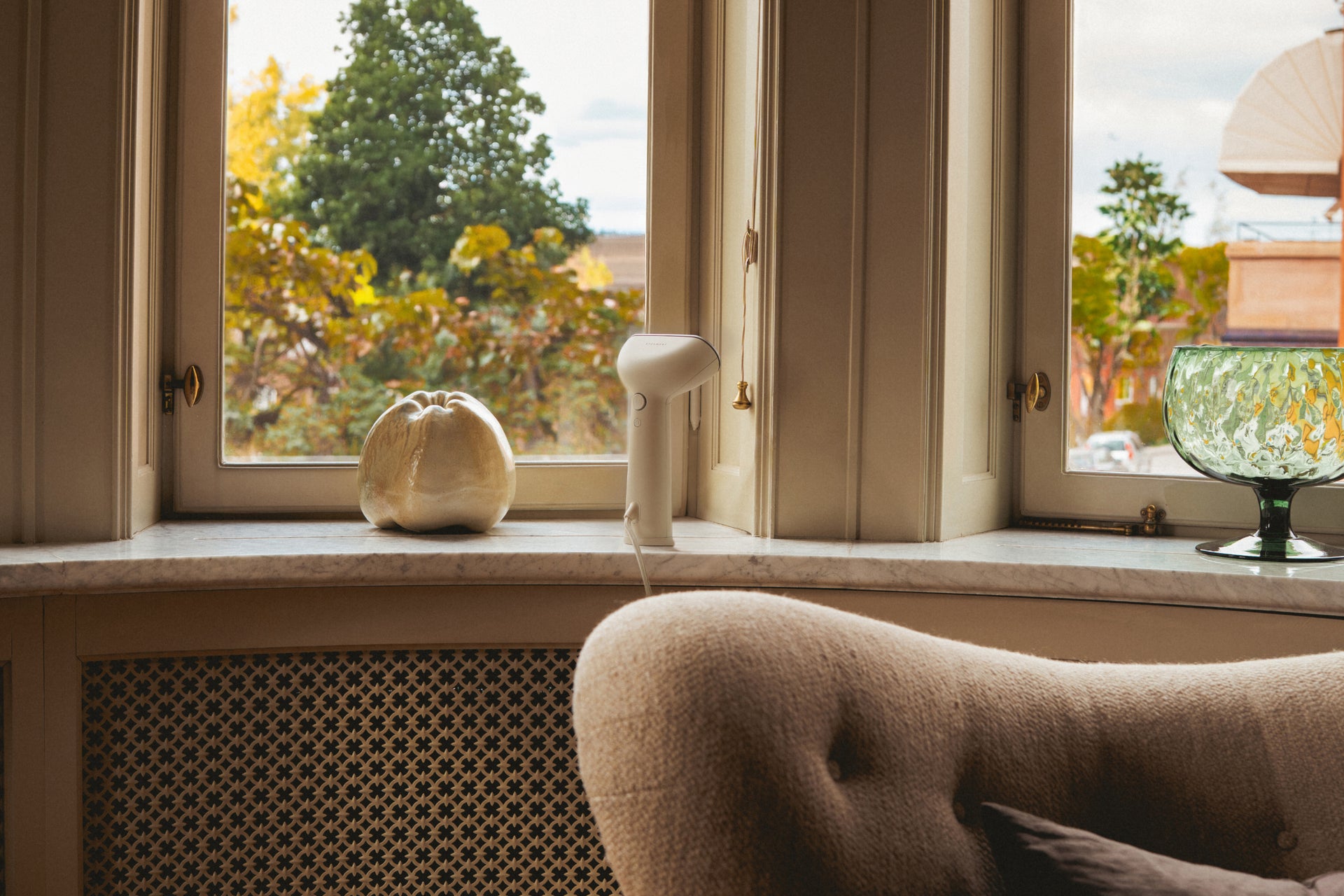 Window with view of trees and a chair in front and a Sand coloured handheld steamer placed by the window