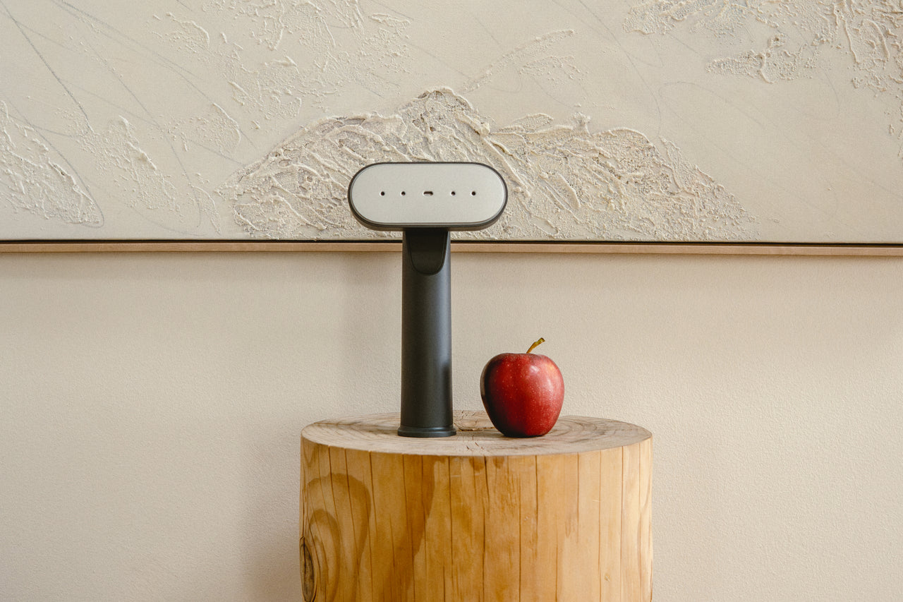 Handheld steamer on a wooden block with an apple next to it against a textured wall.