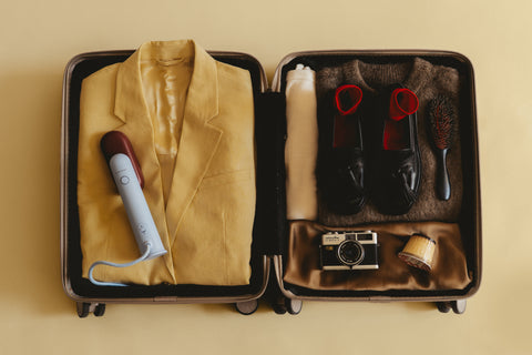 Open suitcase with a yellow jacket, camera a travel steamer and grooming items on a beige background